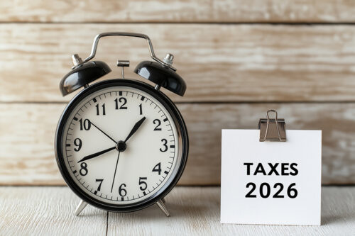Alarm clock and note card displaying TAXES 2026 on a wooden background, symbolizing tax season deadline and financial planning